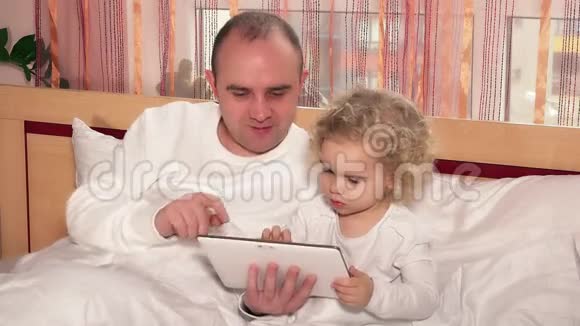 秃头爸爸和小可爱女儿在床上玩平板电脑游戏视频的预览图