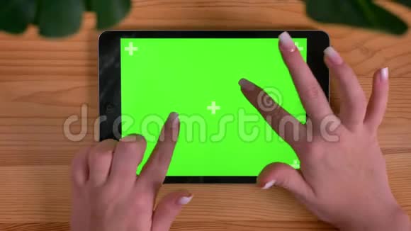 在木制书桌和植物上的水平平板的绿色屏幕上女性双手放大和滑动视频的预览图