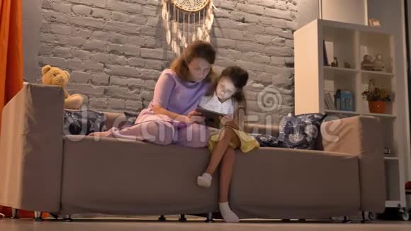 母亲和她的小女儿使用平板电脑坐在客厅的沙发上带着小玩意的家庭概念视频的预览图