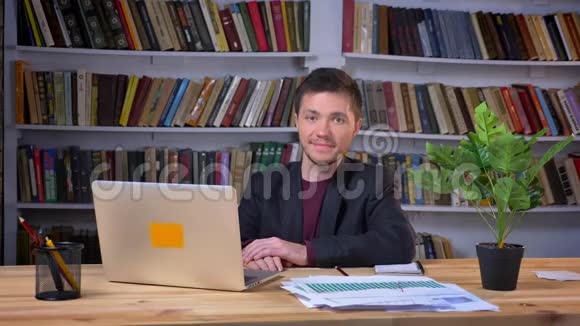 漂亮的成年商人坐在笔记本电脑前看着相机在图书馆里微笑视频的预览图