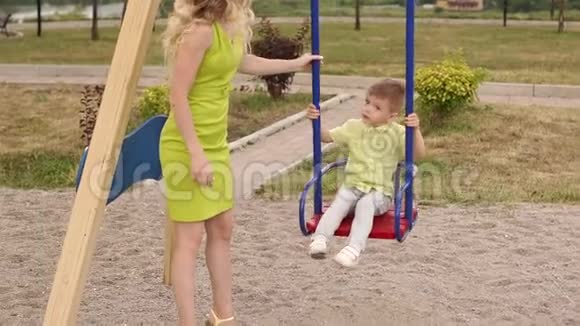 夏天一个漂亮的女人在公园的秋千上滚动她的小儿子视频的预览图