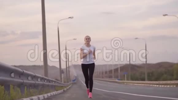 一个穿着白色t恤和轻便运动鞋的运动女孩沿着城外的一条空荡荡的跑道奔跑美丽的森林视频的预览图