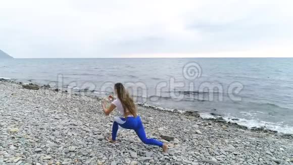 在岩石和海洋背景上做深蹲运动的女孩在呼吸练习时猛击身体视频的预览图