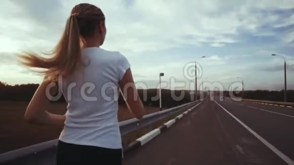 一个穿着白色t恤和长发的运动女孩沿着城外的一条空跑道跑着田径班视频的预览图