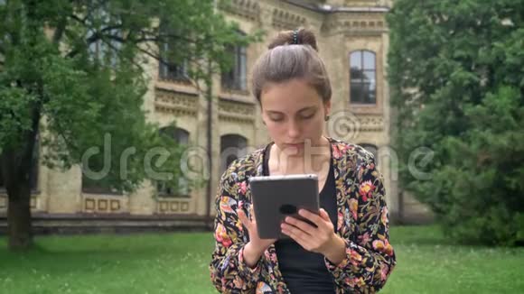 年轻漂亮的女人穿着五颜六色的夹克在平板电脑上浏览坐在大学校园附近的公园里迷人而迷人视频的预览图