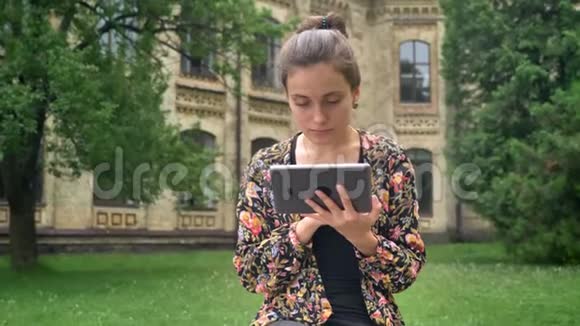 年轻的美女在平板电脑上打字坐在公园附近大学的长凳上拿着草对着镜头微笑视频的预览图