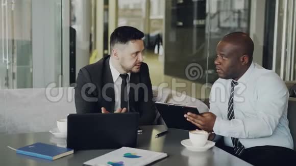 两位商务同事讨论他们的商务合同并在时尚咖啡馆里看记事本非裔美国人视频的预览图