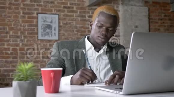 成功的非裔美国金发商人从笔记本电脑上写笔记喝咖啡坐在现代办公室视频的预览图