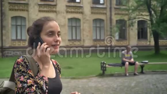 年轻开朗的女人穿着五颜六色的夹克上大学打电话走在大学附近的街道上视频的预览图