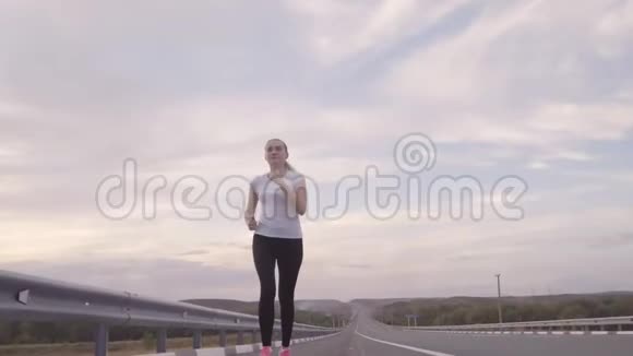 女孩穿着白色的t恤和明亮的运动鞋在日落时在路上奔跑头发在风中飘扬关于视频的预览图