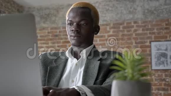 年轻的金发非洲商人在笔记本电脑上打字看着相机自信而成功坐在办公室里视频的预览图
