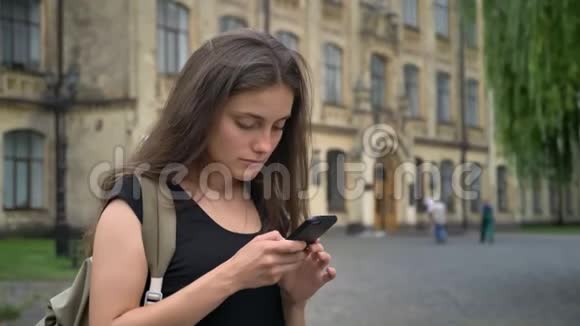 年轻漂亮的白种人学生打手机站在大学附近的公园里女人拿着背包视频的预览图