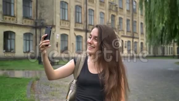白人美女通过手机视频聊天站在街边的大学大楼附近视频的预览图
