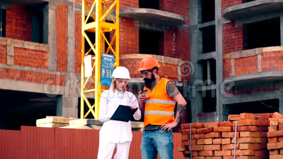 在建筑背景下戴着头盔和文件夹的女人与穿着防护背心的留胡子的男人交流视频的预览图