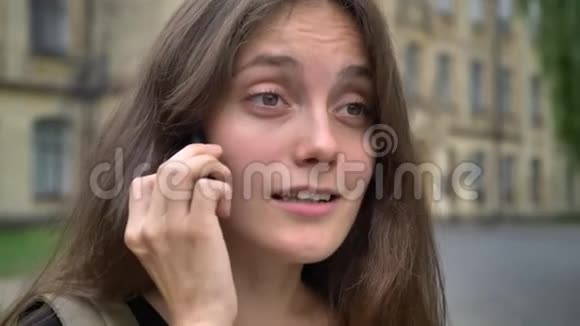 一位留着长发的年轻美女站在大学附近的公园里在电话里谈笑风生视频的预览图