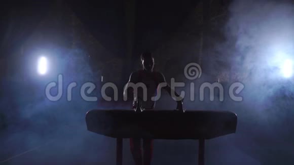 男子体操运动员在黑暗的房间里缓慢地在烟雾中进行鞍马运动体育促进健康和成就视频的预览图