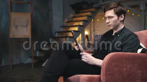 穿着黑色衬衫和牛仔裤的年轻帅哥坐在舒适的大椅子上看着数码平板电脑视频的预览图