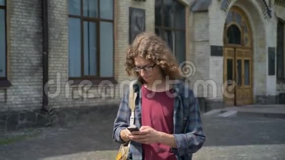 年轻英俊的学生背着背包离开大学打电话发短信走在大学附近的大街上视频的预览图