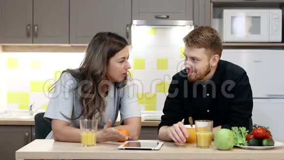 年轻夫妇带着平板电脑坐在厨房的桌子旁吃早餐视频的预览图
