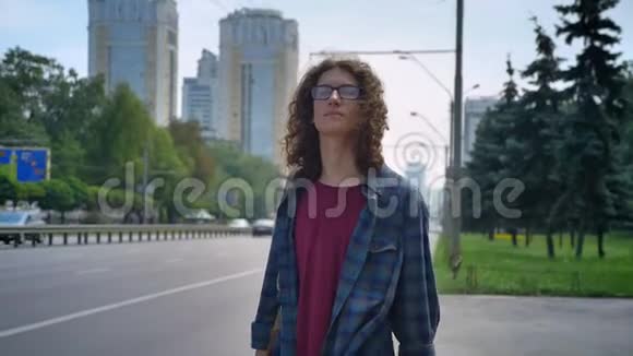 一位戴眼镜的聪明青年留着长长的卷发等着出租车摸着他的头发站在路边的公共汽车站上视频的预览图