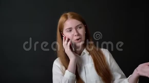 年轻迷人的女人留着长长的姜发在手机上聊天穿着白色衬衫的女孩被隔离在黑色背景上视频的预览图