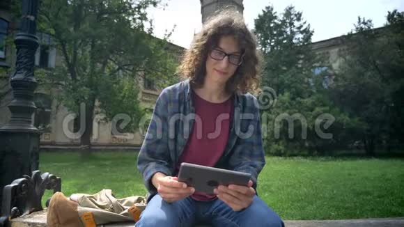 年轻的帅哥戴着一副眼镜卷发拿着平板电脑环顾四周坐在公园附近的长凳上视频的预览图