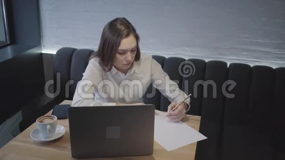 年轻女子用她的笔记本电脑坐在咖啡馆的桌子旁坐在沙发上的女孩拿着一杯咖啡和电脑视频的预览图