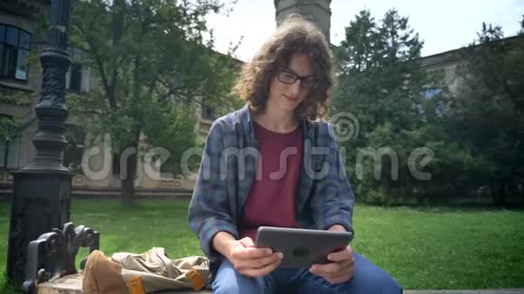 年轻的帅哥一头卷发戴着眼镜拿着平板电脑看着相机坐在公园附近的长凳上视频的预览图