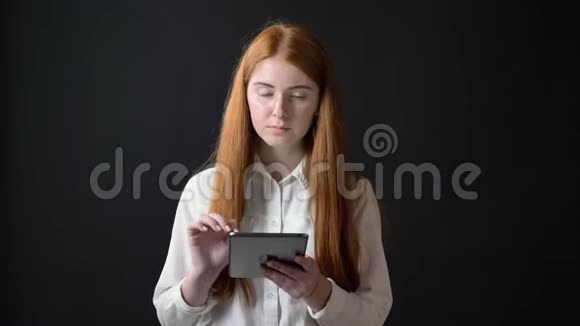 年轻的生姜少女穿着白色衬衫在平板电脑上打字看着相机站在黑色背景上孤立无援视频的预览图