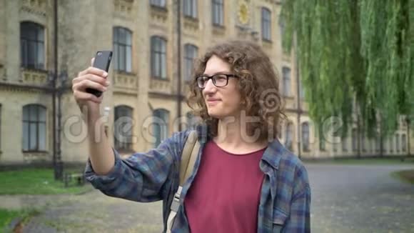 年轻帅气的学生戴着眼镜留着长长的卷发在大学附近的大街上进行视频聊天站在户外视频的预览图