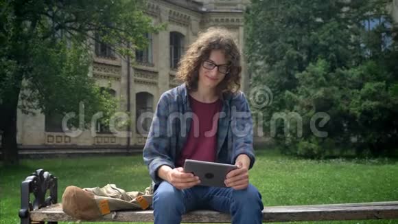 年轻快乐的学生留着长长的卷发在平板电脑上打字坐在大学附近公园的长凳上面带微笑视频的预览图