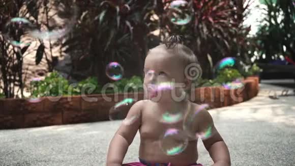 肖像婴儿一岁女孩坐着奶嘴看着花园里的街道上的肥皂泡慢慢地视频的预览图