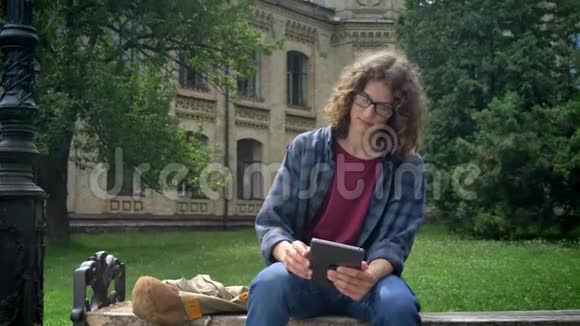 聪明认真的学生留着平板眼镜留着长长的卷发坐在大学或校园附近公园的长椅上视频的预览图