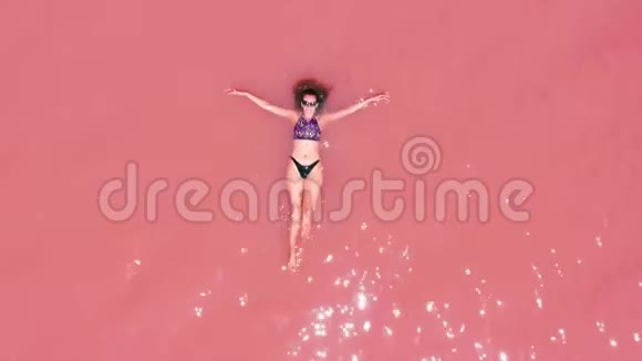 空中观景一个美丽的年轻女孩躺在粉红色的湖里一件比基尼太阳镜粉红色的清澈的水一张最高的风景视频的预览图