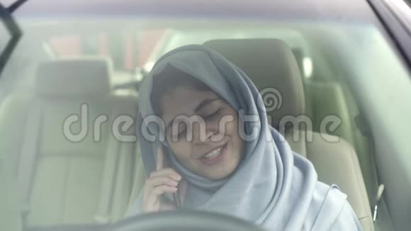 戴着头巾的漂亮年轻女子在车里的智能手机上谈笑风生视频的预览图