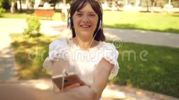 戴着耳机的女孩微笑着带着电话跳舞在公园里和她心爱的男人牵手旋转团队合作视频的预览图