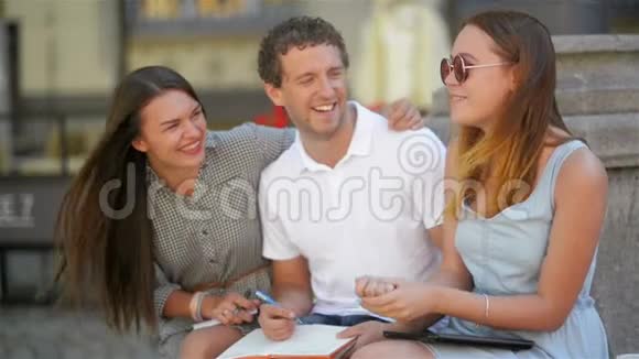 三位积极微笑的青年学生坐在城市公园的壕沟上准备考试的特写镜头视频的预览图