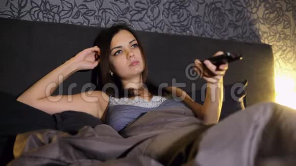 年轻女子在晚上坐在床上看电视时用遥控器换频道瀑布视频的预览图