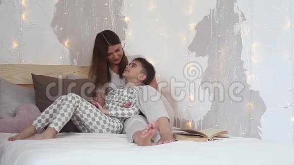 美丽的母亲坐在床上亲吻她的孩子孩子躺在母亲腿上视频的预览图