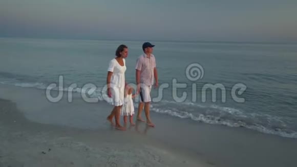 家人在海滩上散步父母和他们的小女儿沿着海滨散步父母蹲在他们身边视频的预览图