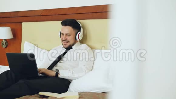 快乐的商人戴着耳机在笔记本电脑上工作在酒店房间里躺在床上时微笑着听音乐视频的预览图
