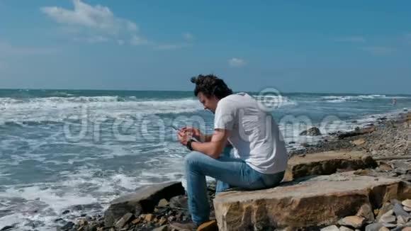 年轻人在坐在海边的大石头上的手机上输入一条信息视频的预览图