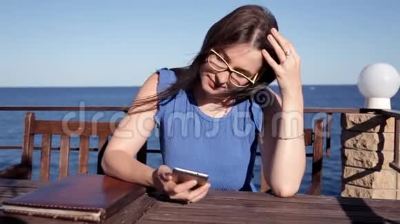 女孩坐在露台上的一家餐馆里俯瞰大海风吹起她的头发打电话视频的预览图