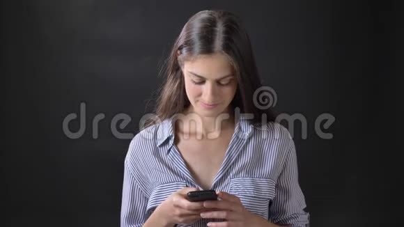 穿着条纹衬衫的漂亮年轻女子在电话上发短信微笑着站在黑色背景上孤立无援视频的预览图