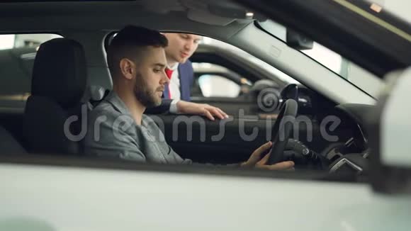 自信的男买车者坐在现代展厅内新车的驾驶座上与推销员交谈视频的预览图