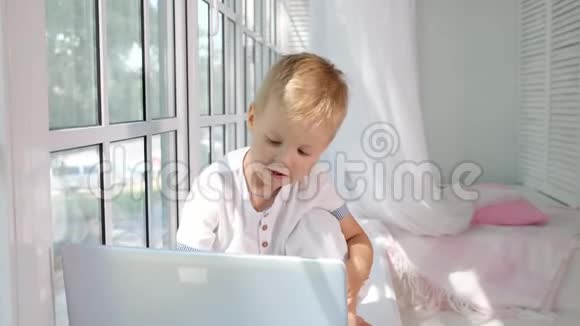 可爱的小男孩坐在大白窗的窗台上看着一台笔记本电脑视频的预览图
