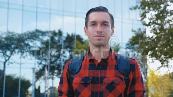 穿格子衬衫和背包看相机的英俊年轻人的肖像大玻璃城市建筑视频的预览图