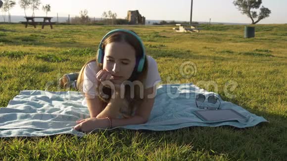 女人躺在公园的草地上用耳机听音乐视频的预览图