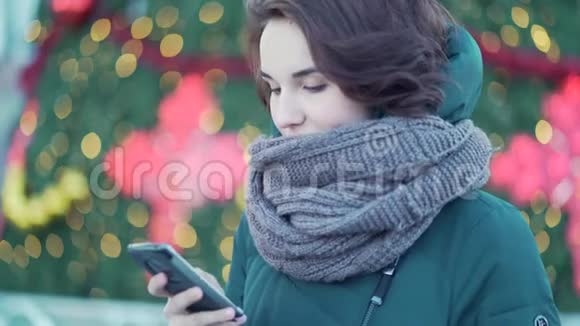穿着绿色冬季夹克的女孩在她的智能手机上打字站在装饰好的圣诞树附近特写镜头视频的预览图