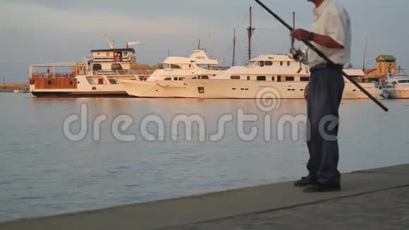 人钓鱼用鱼竿站在码头上背景是美丽的白色游艇渔夫转身视频的预览图